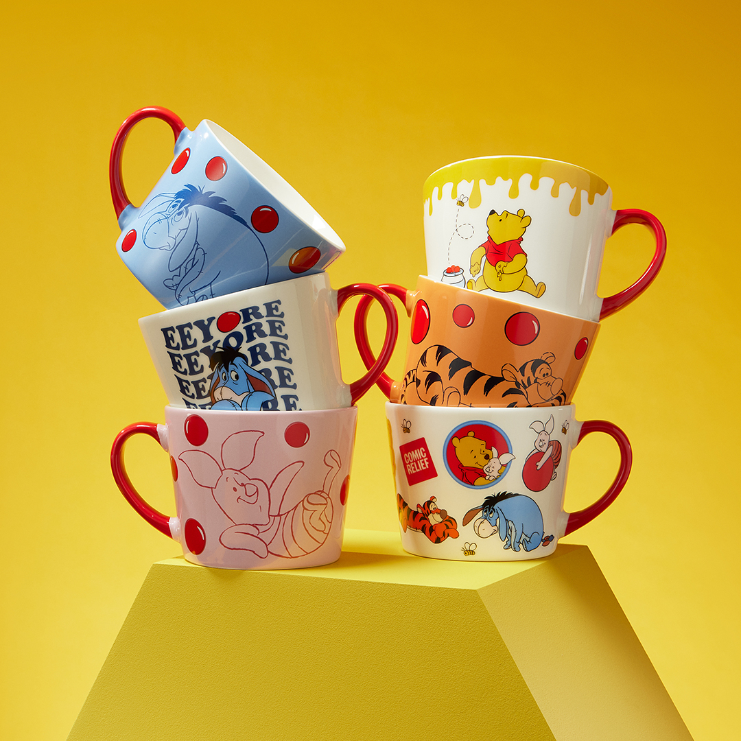 A stack of six colourful Winnie the Pooh–themed mugs arranged in two leaning columns against a bright yellow background. Each mug features different character illustrations, including Pooh, Piglet, Tigger, and Eeyore, along with playful red balloon motifs and honey-inspired details.